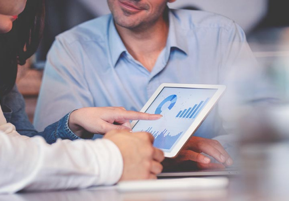 Business people working on financial data on a digital tablet. One looks like the manager working with employees, or a financial advisor. It may also be a training session. All are dressed in business casual clothing.