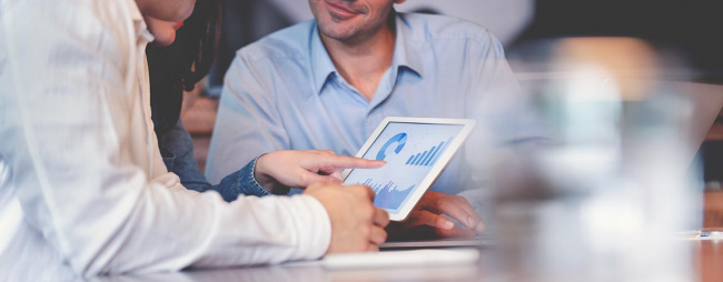 Business people working on financial data on a digital tablet. One looks like the manager working with employees, or a financial advisor. It may also be a training session. All are dressed in business casual clothing.